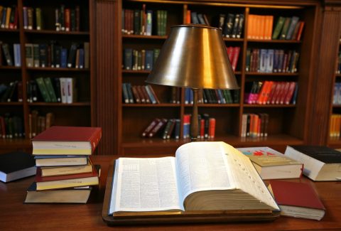 Open book on a wooden library desk with a brass reading lamp, surrounded by stacked books.