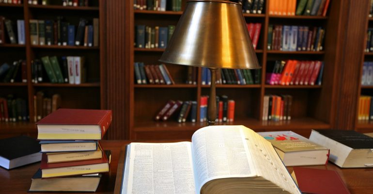 Open book on a wooden library desk with a brass reading lamp, surrounded by stacked books.
