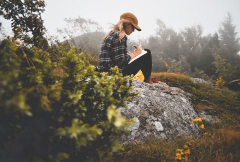 A woman sits on a rock writing in a journal, reflecting themes of self-reflection, emotional wellbeing, and healing through reading and writing.