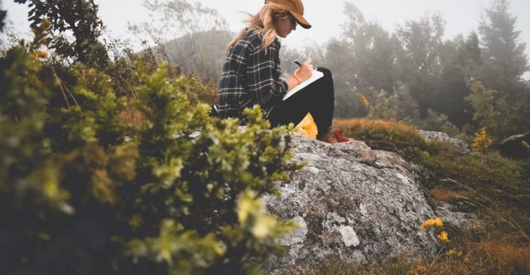 A woman sits on a rock writing in a journal, reflecting themes of self-reflection, emotional wellbeing, and healing through reading and writing.